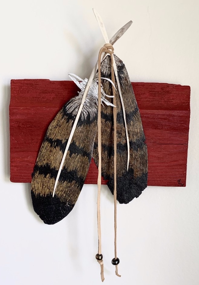 Two feathers connected with deer hide mounted on a a piece of red reclaimed barn wood