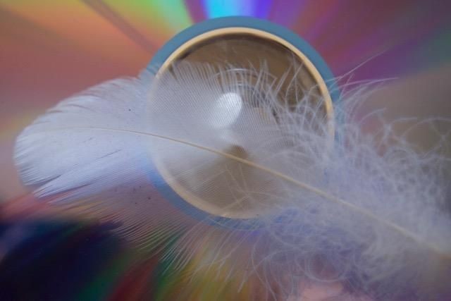 white feather in front of a blue circle with glass in it. the background is a rainbow